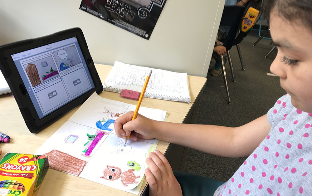 A young student is seated at a desk, working on a colorful drawing for a fractured fairy tale project. The student is using a pencil to add details to the illustration on paper, which shows vibrant scenes featuring animals. An iPad is positioned in front of the student, displaying a digital comic panel on the Book Creator app. Crayons and other drawing tools are spread out on the desk, indicating a blend of digital and traditional art techniques.